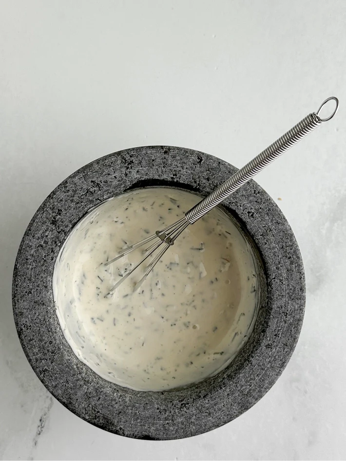 Photograph taken from above of a molcajete filled with the mixed rosemary tarragon aioli and a small metal whisk