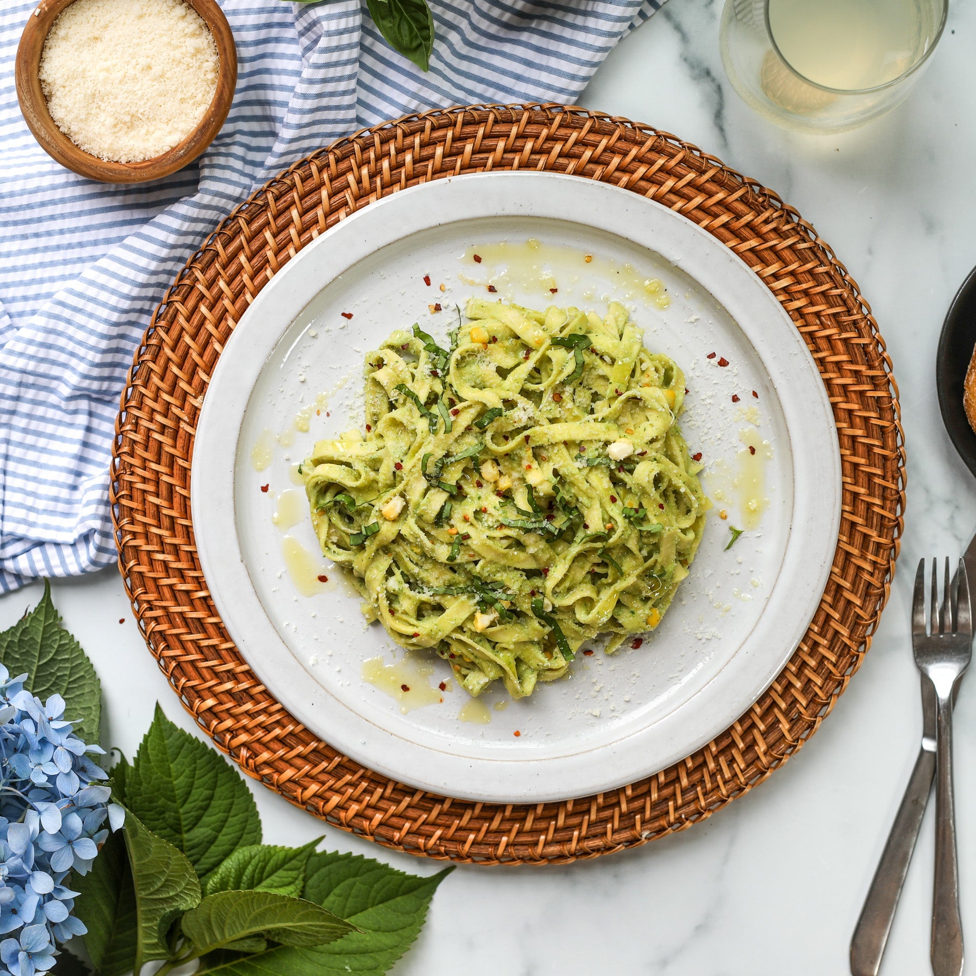 A plate of Corn & Basil Cream Pasta. Pasta is on a white plate atop a woven, wooden charger on white marble. To the left of the plate is a blue-and-white-striped  towel and  blue hydrangeas. To the right is a bowl of sliced bread and a fork and knife.