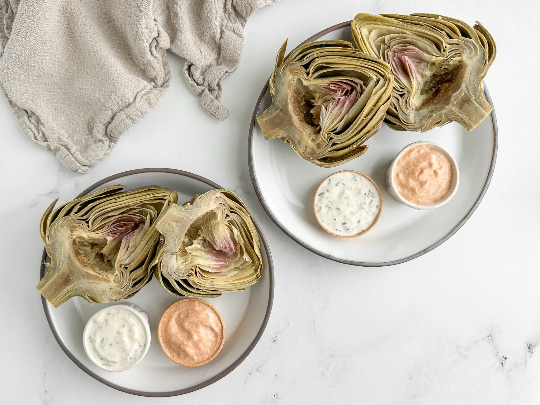 Photograph of two identical white ceramic plates with a  roasted artichoke that has been cut in half and is served next to a small bowl of rosemary tarragon aioli and  another small bowl of garlic lemon mitmita aioli.