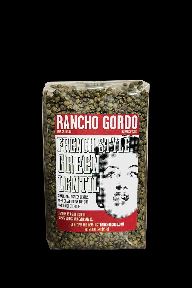A One Pound Bag of Rancho Gordo French-Style Green Lentils on a white background. Red and white label with an image of the face of a woman licking her upper lip. Additional text on bag reads: Small, meaty green lentils. West Coast-grown for our own unique terroir. Famous as a side dish, in stews, soups, and even salads.