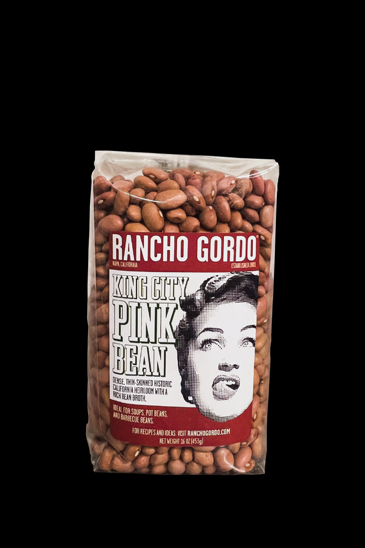 A One Pound Bag Of Rancho Gordo  King City Pink Beans on a white background. Red and white label with an image of the face of a woman licking her upper lip. Additional text on bag reads: Dense, thin-skinned historic California heirloom with a rich bean broth. Ideal for soups, pot beans, and barbecue beans.