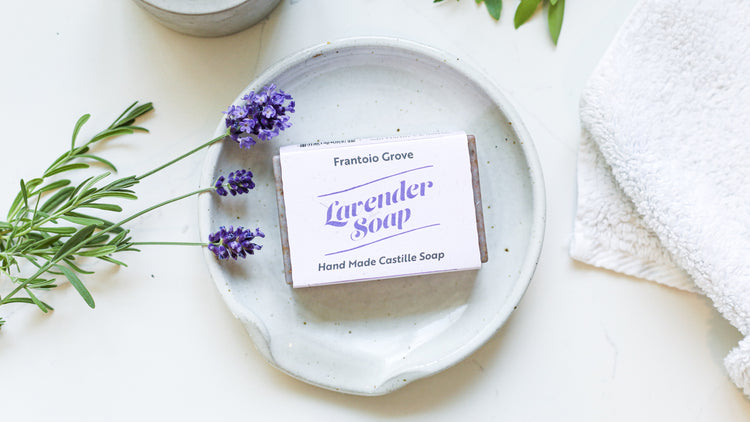 Photograph of a bar of Frantoio Grove Lavender soap on a white earthenware plate. Also in the picture are three sprigs of fresh lavender and a white hand towel.