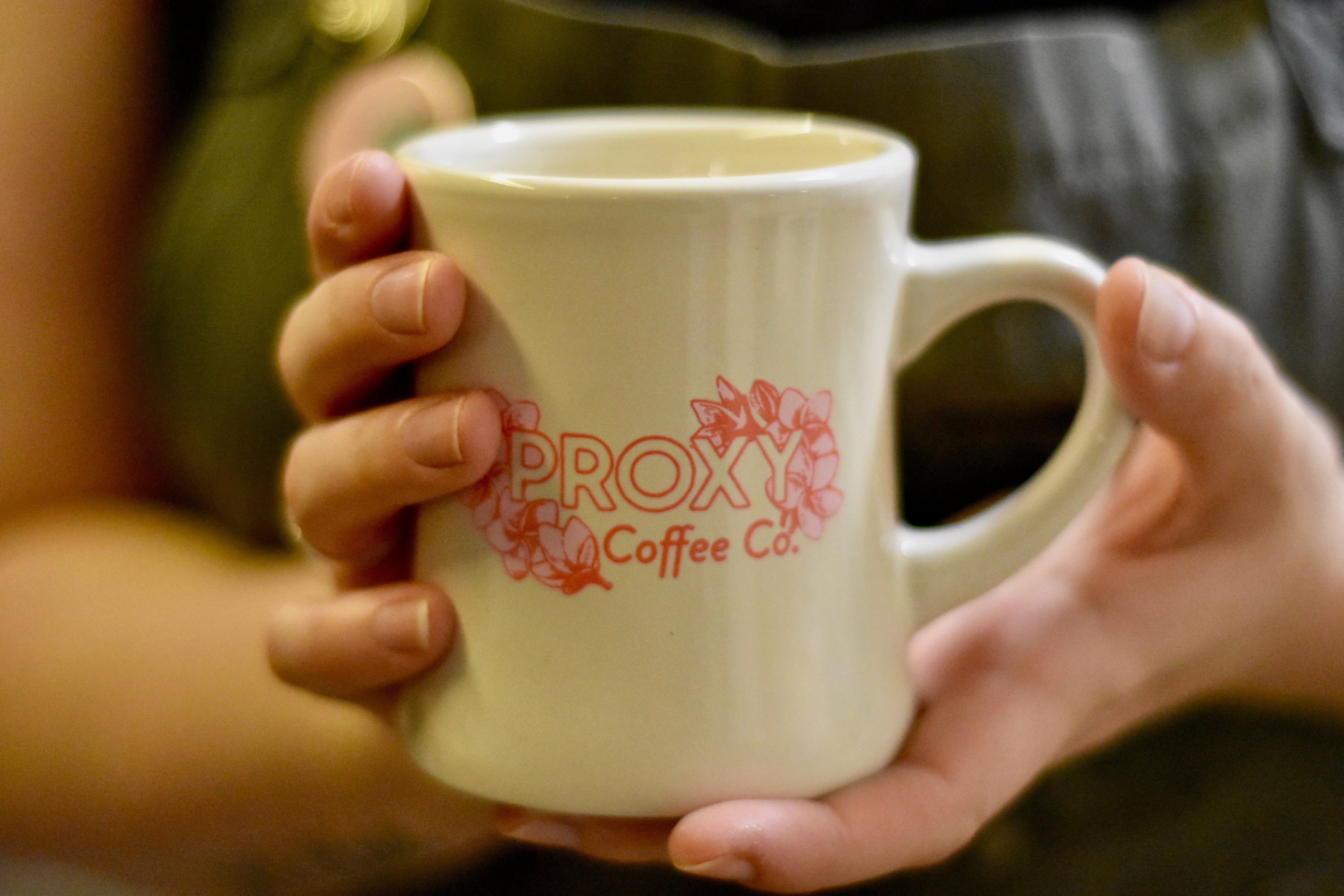Close up photograph of a person's hands holding a white, diner-style coffee mug with the words "Proxy Coffee Co." and images of plumeria flowers printed on the mug in pink.
