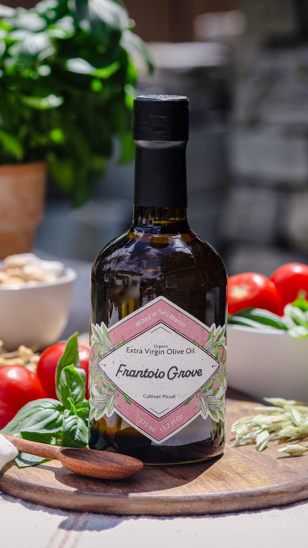 Photograph of a bottle of Frantoio Grove Picual EVOO sitting on a round wooden cutting board. Also on the cutting board is a wooden spoon, a sprig of fresh basil, and a dried grain. On the table behind the bottle is a bowl of tomatoes and fresh basil, another bowl of garlic, and a terracotta pot with fresh basil growing it.