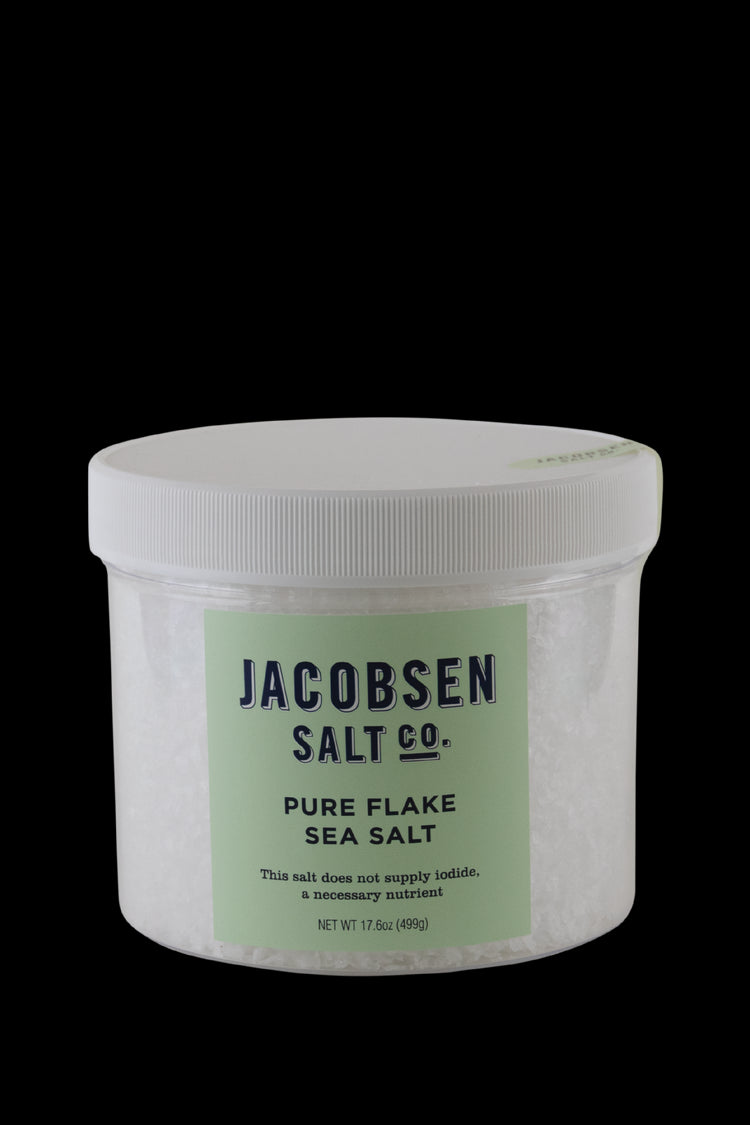 A Large Plastic Jar Of Jacobsen Salt Co's Pure Flake Sea Salt With Celeste Green Label and White Background. Label says "This salt does not supply iodide, a necessary nutrient."