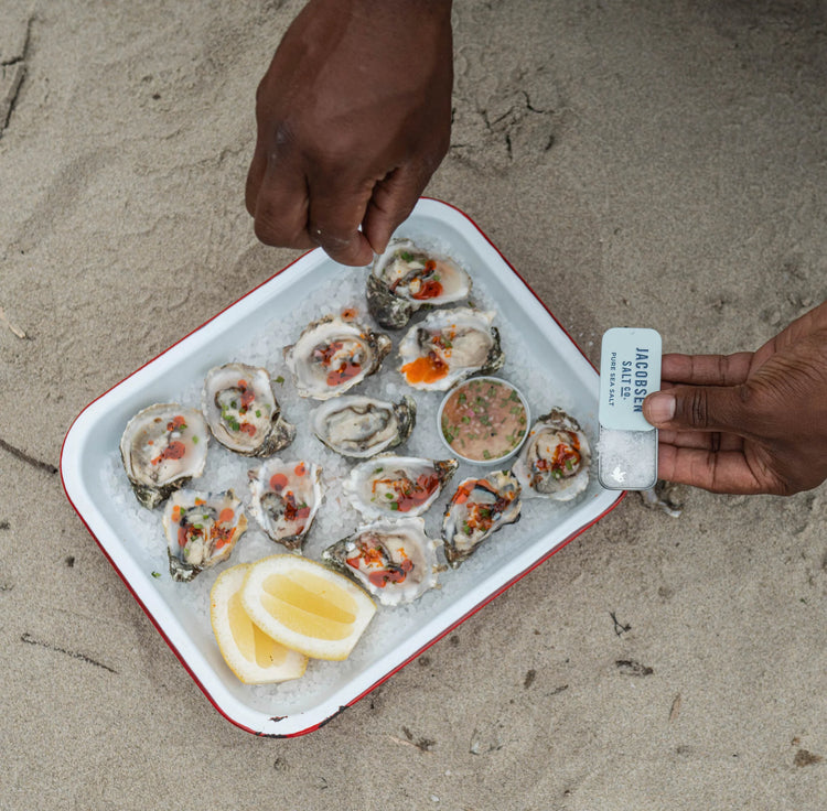 A Photo of a tray of raw oysters with hot sauce and lemon wedges sitting on the sand. Close shot of hands holding a Jacobsen Salt Co. slide tin of pure flake salt and sprinkle a pinch onto the oysters.