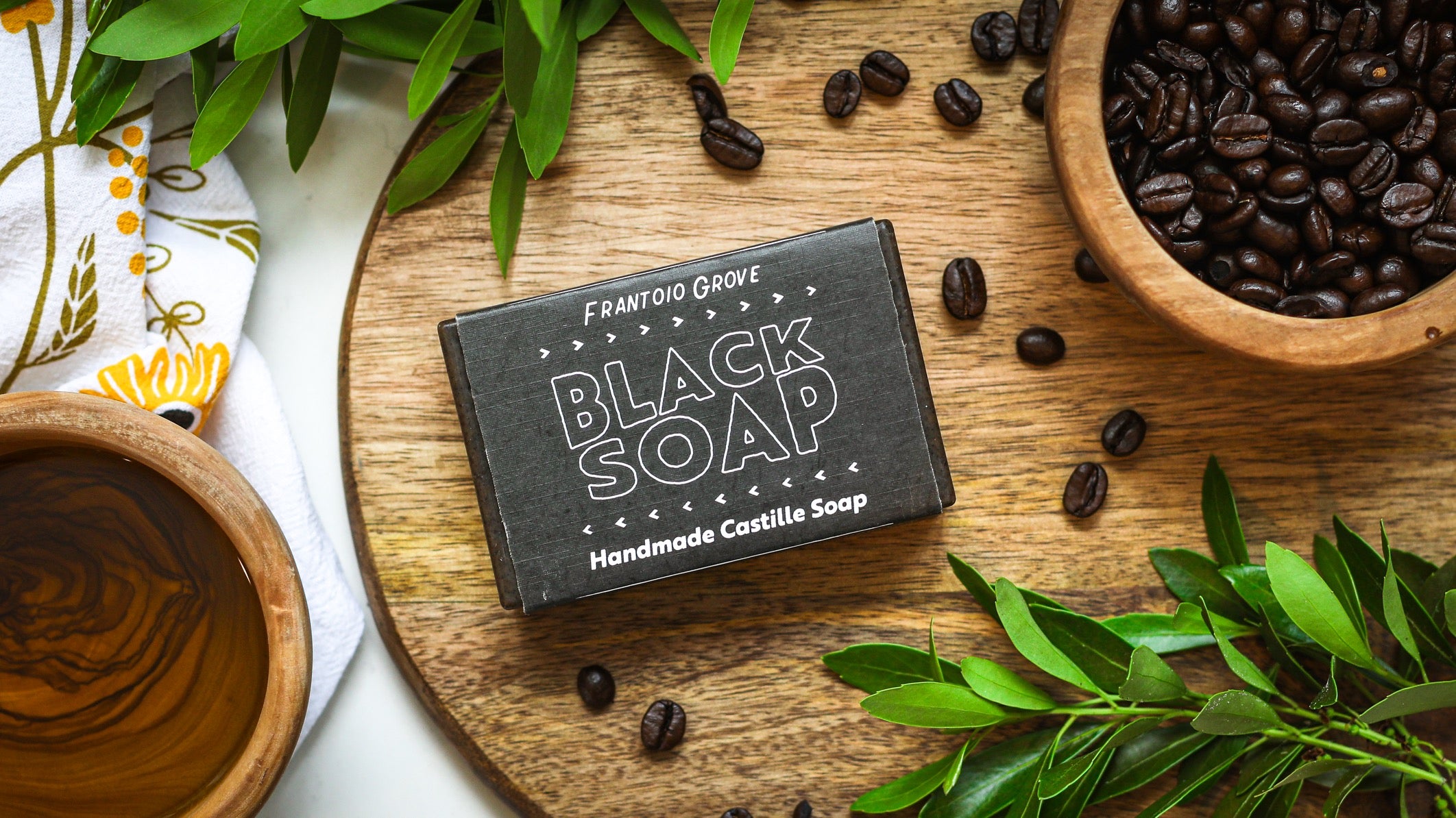 Photograph of a bar of Frantoio Grove Black Soap on a light wooden cutting board. Also on the board are some loose, scattered coffee beans, a wooden bowl filled with coffee beans, and some bright green leafy branches. Next to the cutting board is an empty wooden bowl and a white tea towel with an abstract image of a sunflower.