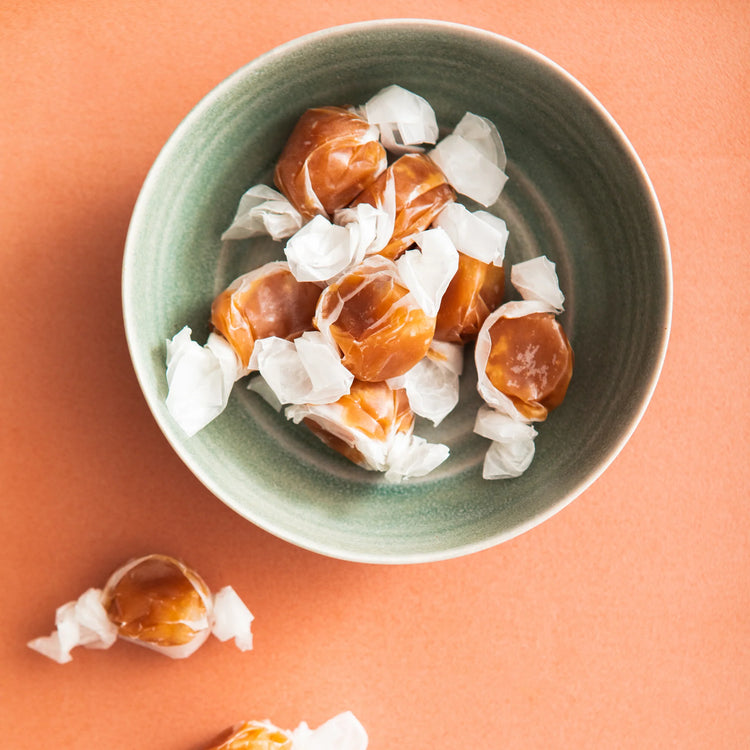 A Photo of a pale green bowl of caramels in their wax paper wrappers on an orange background.