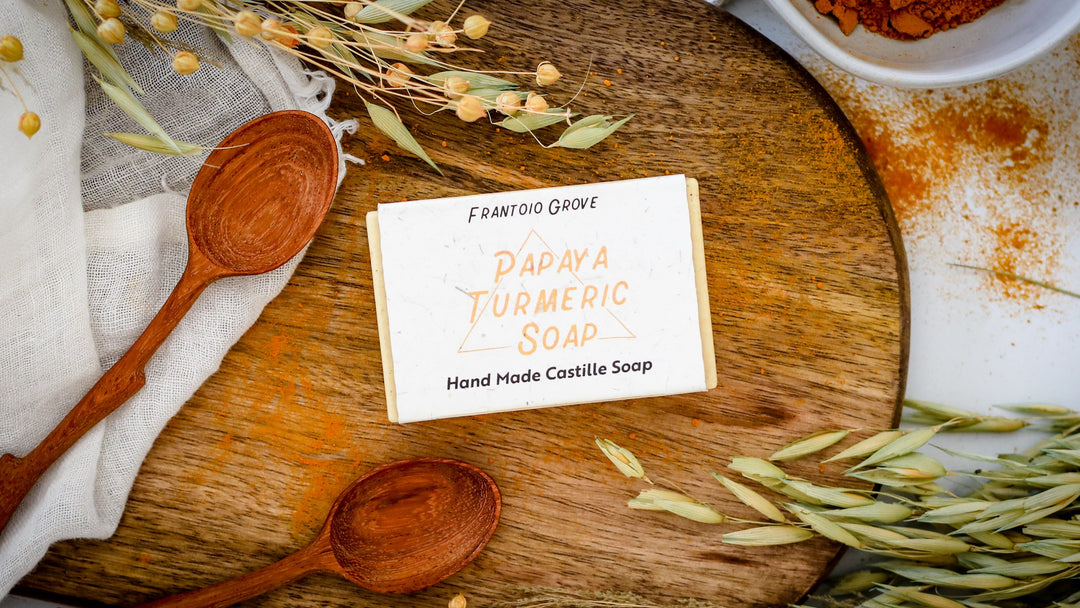 Photograph of a bar of Frantoio Grove Papaya Turmeric Soap on a wooden cutting board. Also on the cutting board are two reddish wooden spoons, a thin cotton tea towel, some dried grains, and a sprinkling of ground turmeric. Part of a white bowl full of ground turmeric is in the upper right-hand corner of the picture.