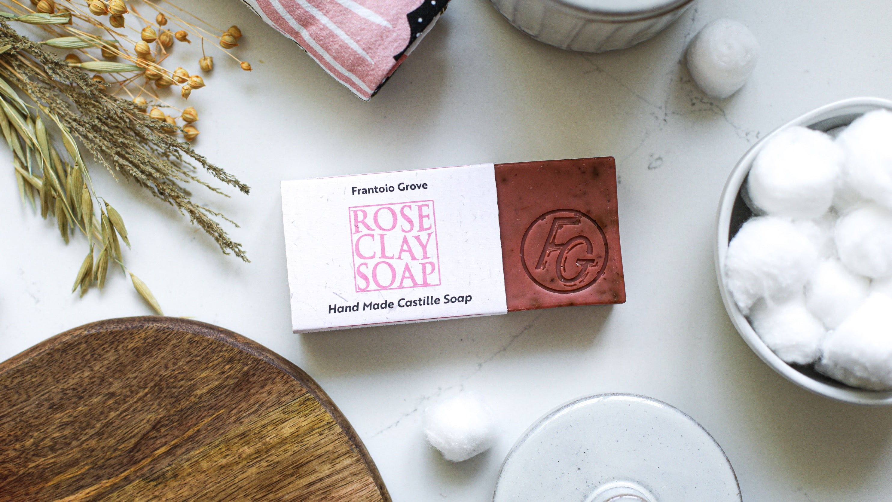 Photograph of a bar of Frantoio Grove Rose Clay Soap on a white marble surface. The soap sticks out from its wrapper to show "FG" with a circle around it stamped into the bar. Also in the picture are the edge of a wooden cutting board, a white container filled with cotton balls, the edge of a pink, black, and white tea towel, some dried grains, and two loose cotton balls.