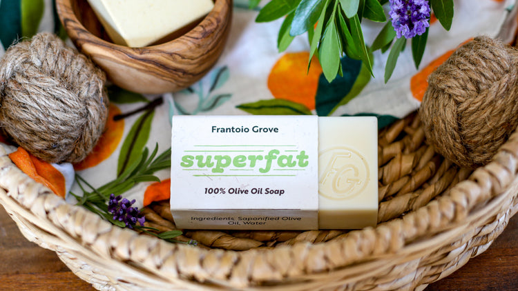 Photograph of a bar of Frantoio Grove Superfat Soap sitting inside a woven basket on top of a white tea towel with a print of oranges. The soap sticks out from its wrapper to show "FG" with a circle around it stamped into the bar. Also in the basket are two balls of rough twine, some sprigs of lavender and greenery, and a wooden bowl with another bar of soap in it.