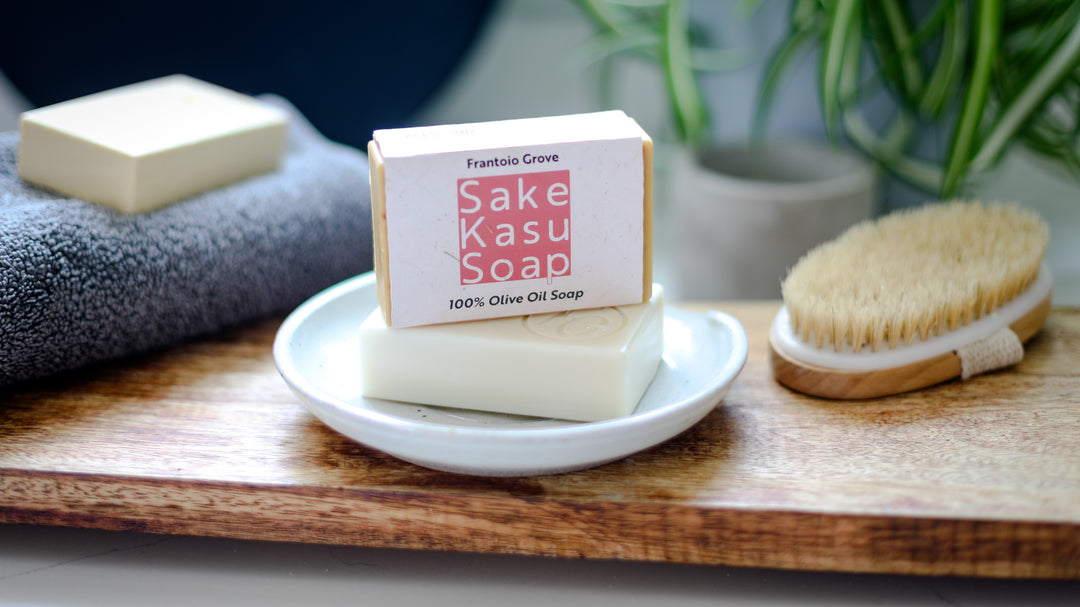 Photograph of a bar of Frantoio Grove Sake Kasu Soap, sitting on top of another bar of Frantoio Grove soap in a white ceramic dish. Dish is sitting on a wooden surface next to a boar bristle dry-brush on the right side and another bar of soap sitting on a folded grey bath towel on the left. In the background, slightly out of focus, is an empty grey pot and a spider plant.