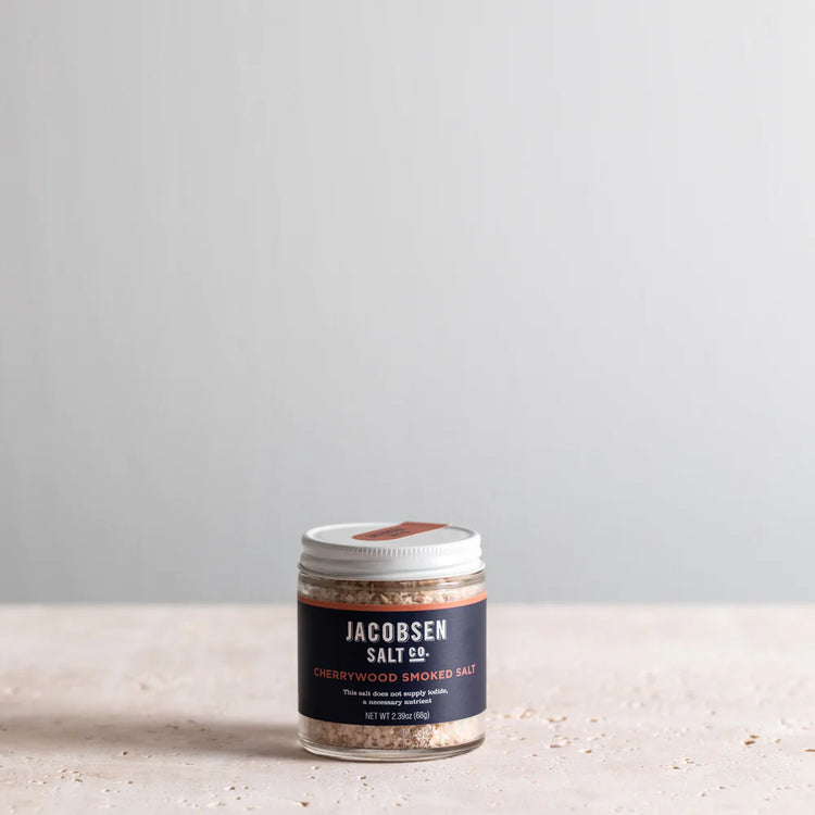 A Styleized Photo of |Jacobsen Salt Co's Cherrywood Smoked Salt With Blue Label. It is shot on a stone surface with soft lighting 