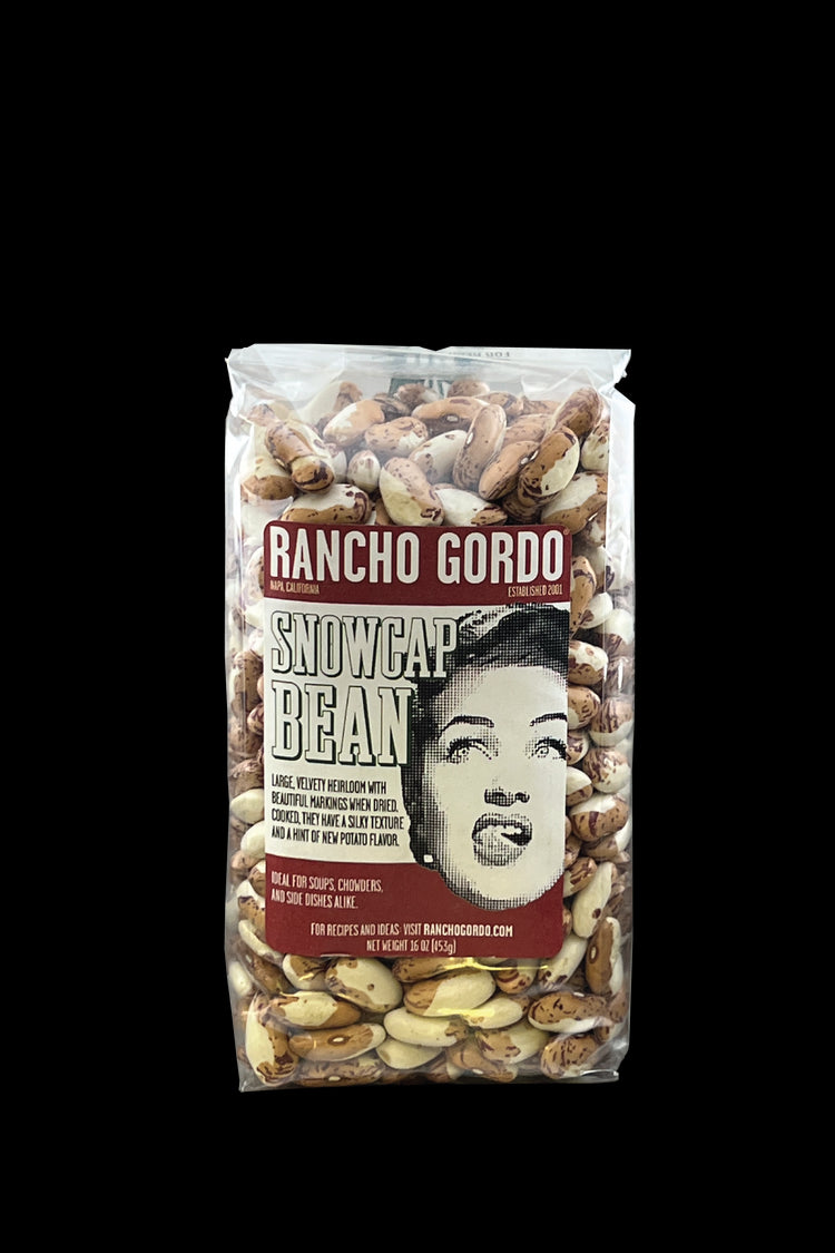A One Pound Bag Of Rancho Gordo Snowcap beans on a white background. Red and white label with an image of the face of a woman licking her upper lip. Additional text on bag reads: Large, velvety heirloom with beautiful markings when dried. Cooked, they have a silky texture and a hint of new potato flavor. Ideal for soups, chowders, and side dishes alike.